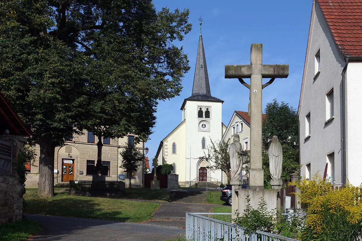 Die Pfarrkirche St. Gallus mit ihrem spitzen Kirchturm und das Wegkreuz davor.